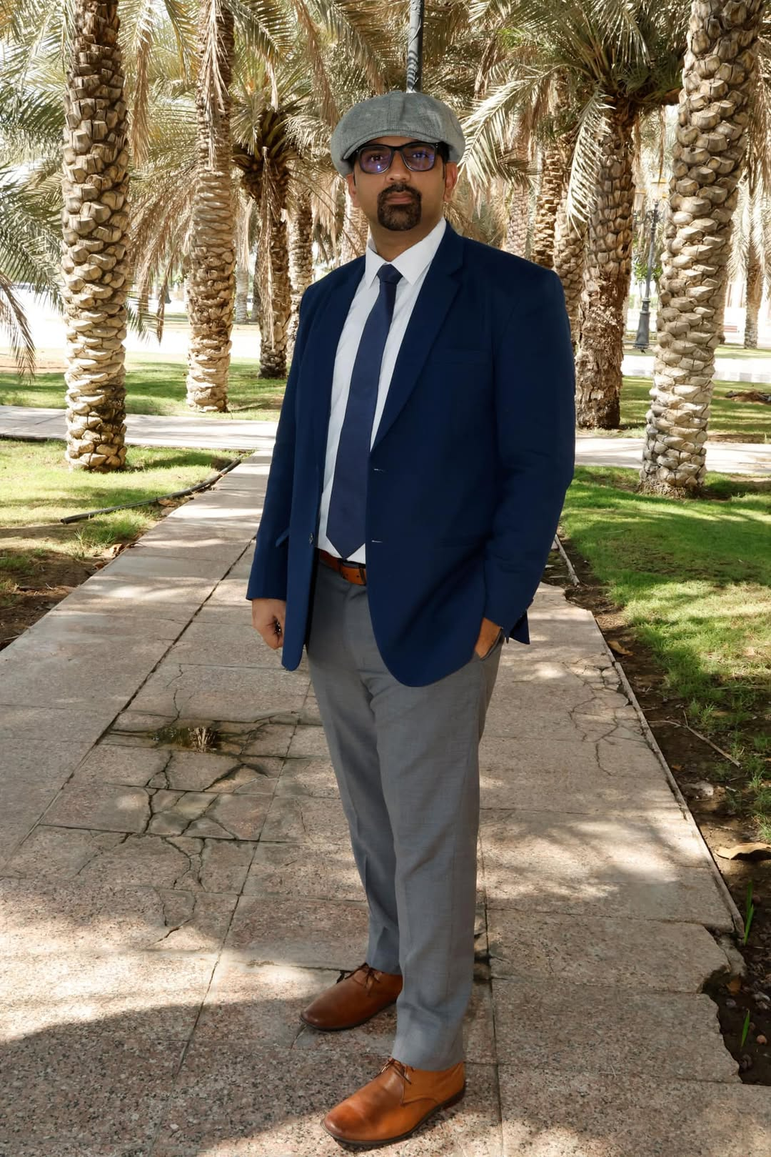 Muhammad Awais wearing a navy suit and grey cap, photographed outdoors among palm trees